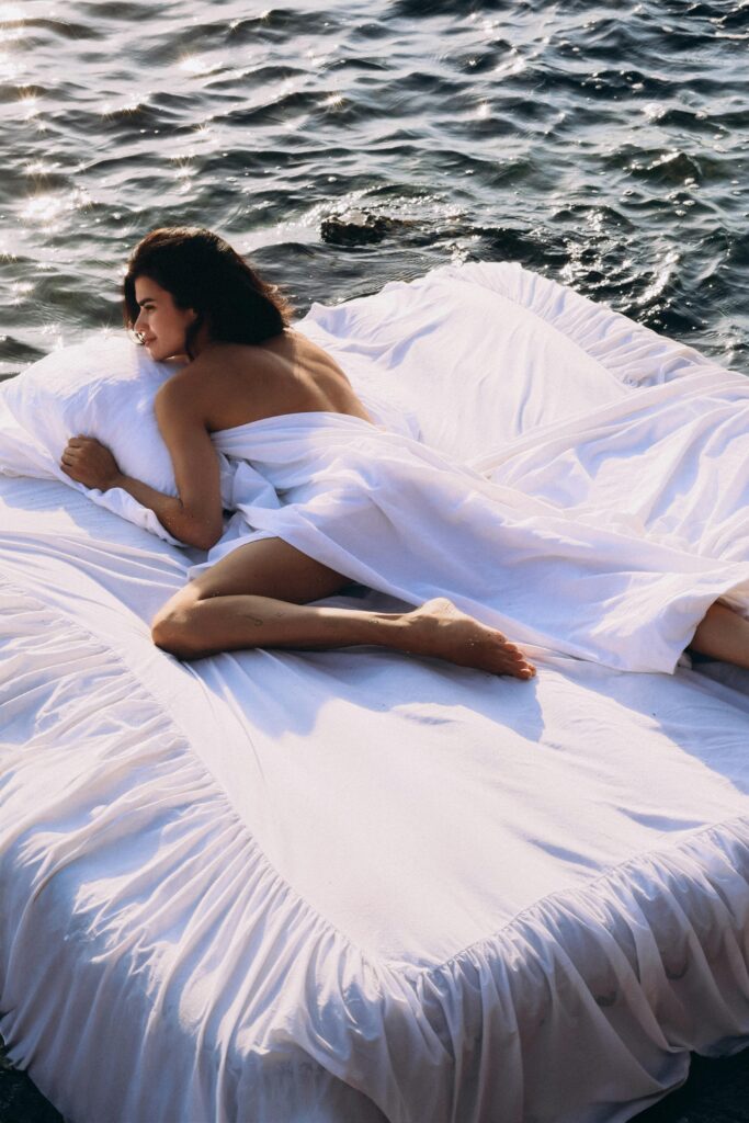 A woman enjoys sunbathing on a floating bed draped in white sheets by the ocean.
